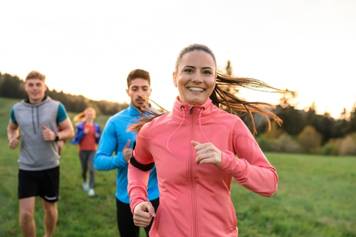 A group of joggers are running through a field. A woman in a pink jacket is in front and smiling.